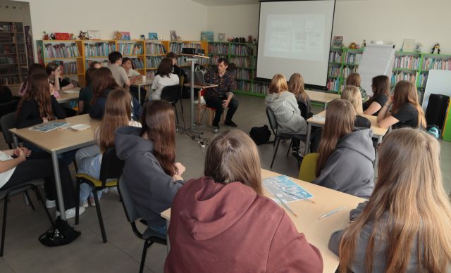 młodzież siedzi przy stolikach, przed nimi przy laptopie siedzi mężczyzna, w tle regały z książkami
