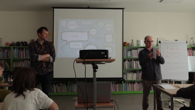 młodzież siedzi przy stolikach, przed nimi stoi dwóch mężczyzn, w tle regały z książkami