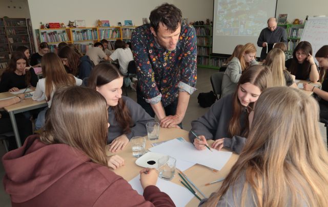 mężczyzna pochylony nad 4 siedzącej młodzieży przy stoliku, na stole katki i pędzle