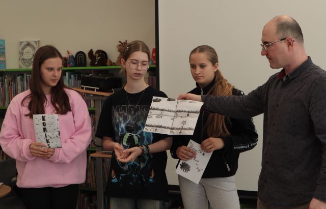 3 dziewczyny stoją i pokazują swój komiks, obok stoi mężczyzna, w tle ekran