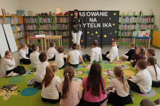 prezdszkolaki siedzą na dywanie, przed nimi stoi dyrektorka biblioteki, w tle regały na książki