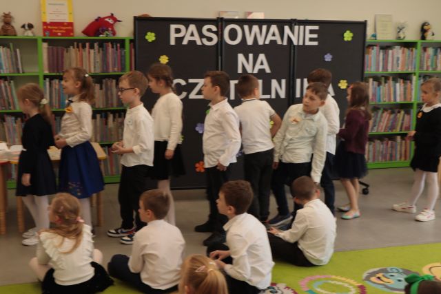 rządek dzieci stoi przed dziećmi siedzącymi na dywanie