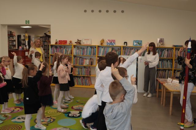 dzieci stoją przed 2 bibliotekarkami z podniesionymi do góry prawymi rękami