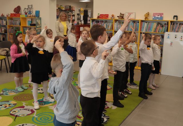 dzieci stoją przed 2 bibliotekarkami z podniesionymi do góry prawymi rękami