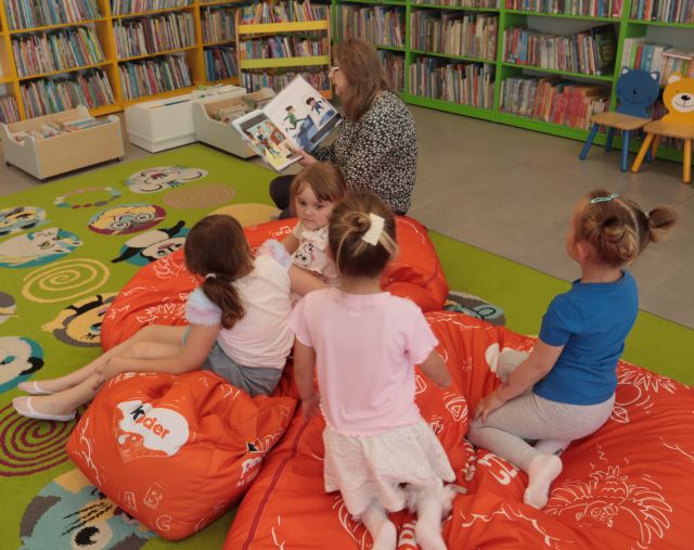 dzieci i bibliotekarka z książką siedzą na czerwonych pufach