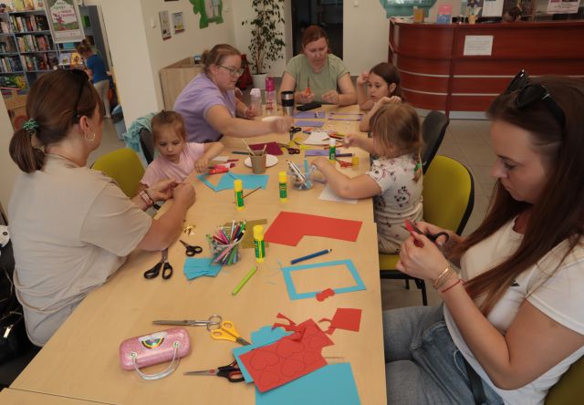 dzieci w rodzicami siedzą przy połączonych stołach, na stołach kolorowe kartki, dzieci i rodzice mają w dłoniach nożyczki