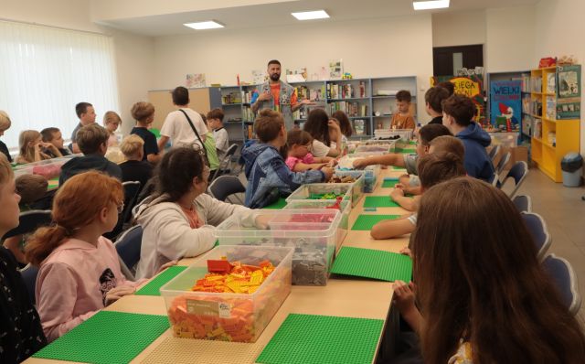 przy stołach z klockami Lego siedzą dzieci, przed nimi stoi uczestnik programu telewizyjnego Lego Masters