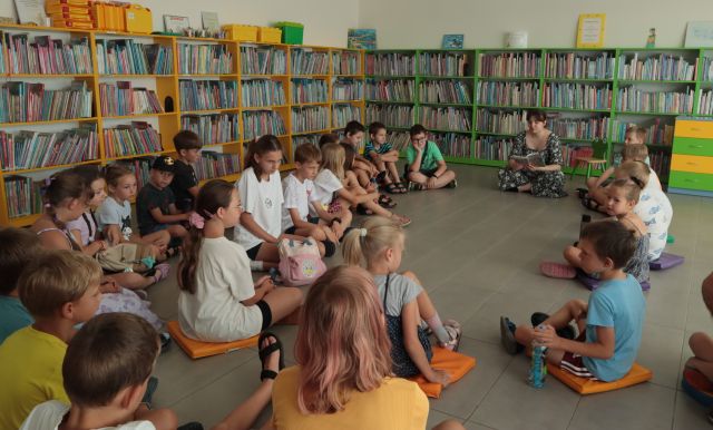 dzieci i bibliotekarka siedzą na pufach w okręgu, w tle regały z książkami
