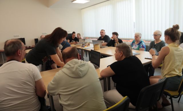 15 dorosłych osób siedzi przy stołach, na stołach laptopy, przy jednym laptopie pochylona dyrektorka biblioteki