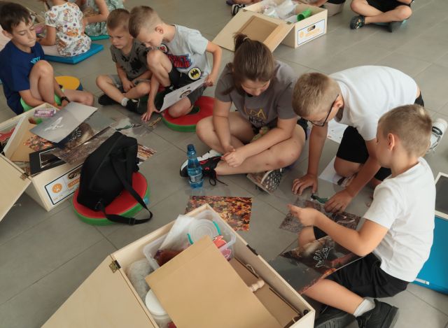 dzieci siedzą na podłodze, przed nimi eduskrzynki z różnymi materiałami