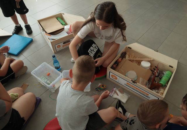 dzieci siedzą na podłodze, przed nimi eduskrzynki z różnymi materiałami
