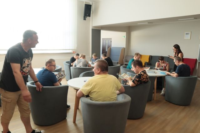 dorosłe osoby siedzą na fotelach przy okrągłych stołach i oglądają komiksy