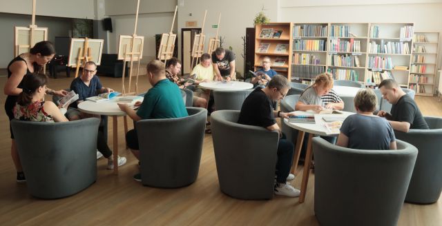dorosłe osoby siedzą na fotelach przy okrągłych stołach i oglądają komiksy