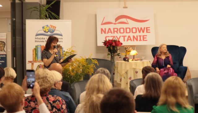 przed publicznością siedzącą na krzesłach stoi dyrektorka biblioteki z mikrofonem