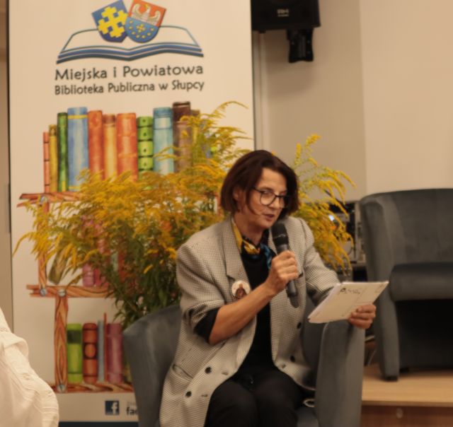 kobieta czytająca z kartki do mikrofonu, za nią rośliny w wazonie