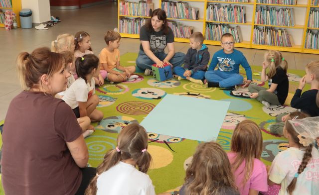 dzieci, nauczycielki i bibliotekarki siedzą na kolorowym dywanie, na śrdoku leży duża niebiska kartka, w tle regały z książkami