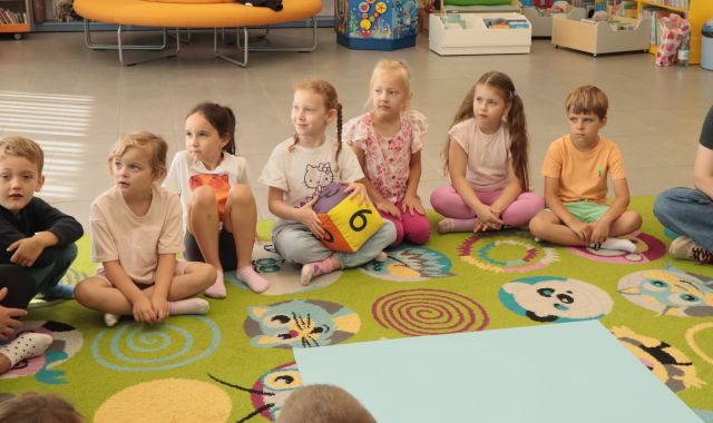 dzieci siedzą na kolorowym dywanie, przed nimi leży duża niebieska kartka, dziewczynka trzyma kolorową dużą kostkę