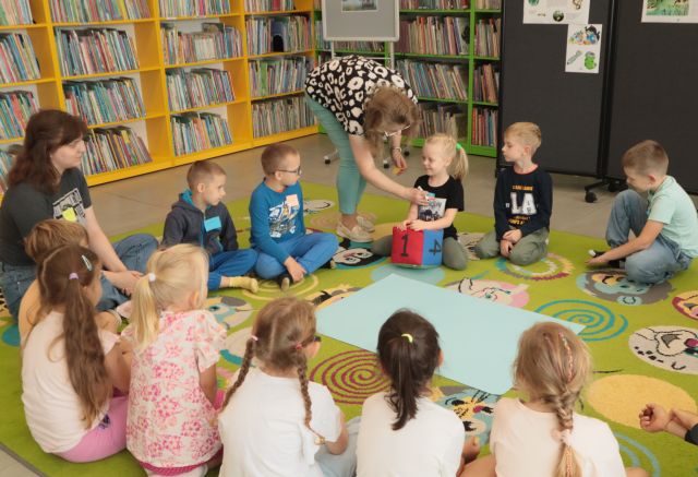 bibliotekarka przypina dzieciom siedzącym na kolorowym dywanie karteczki z imionami