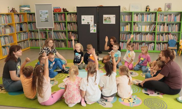 na kolorowym dywanie siedzą dzieci, trzymają w górze dłonie, w tle czarna tablica z plakatami i regały z książkami