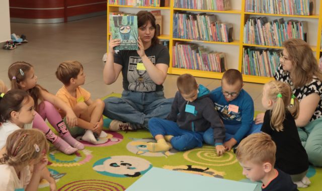 dzieci i bibliotekarka z książką uniesioną do góry siedzą na kolorowym dywanie