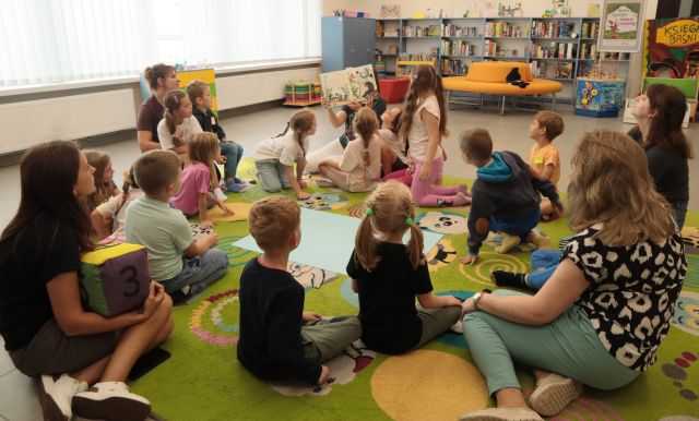 dyrektorka biblioteki pokazuje dzieciom siedzącym wokół niej na kolorowym dywanie ilustracje w książce