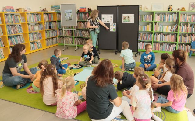 bibliotekarka pokazuje dzieciom siedzącym na dywanie plakaty przypięte do dużej czarnej tablicy