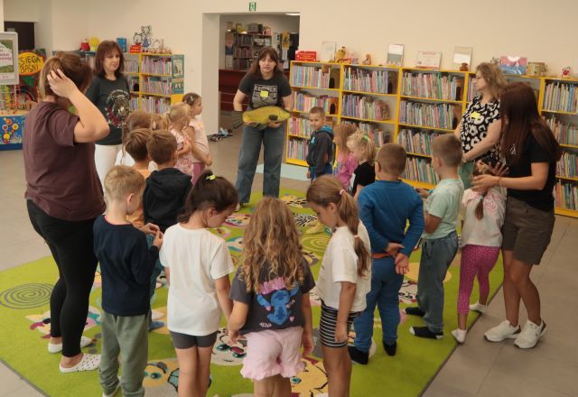 dzieci i bibliotekarka stoją na dywanie, bibliotekarka trzyma dużą maskotkę ryby