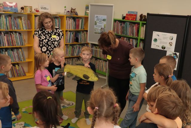 dzieci i dwie kobiety stoją. chłopiec trzyma w rękach maskotkę ryby, w tle regały z książkami