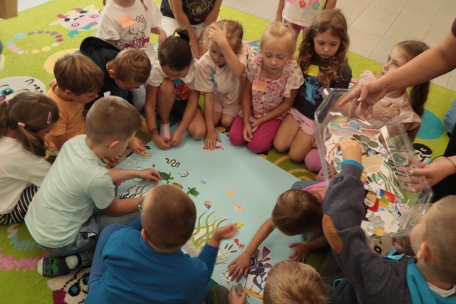 dzieci siedzą wokół dużej niebieskiej kartki, układaja na niej rybki i roślinki i inne zwierzęta wodne