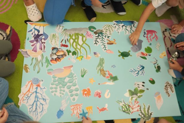 dzieci siedzą wokół dużej niebieskiej kartki, układaja na niej rybki i roślinki i inne zwierzęta wodne