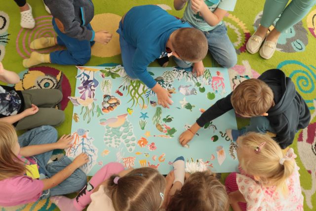 dzieci siedzą wokół dużej niebieskiej kartki, układaja na niej rybki i roślinki i inne zwierzęta wodne