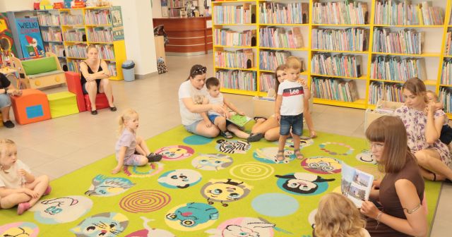 dzieci z rodzicami siedzą na kolorowym dywanie wokół bibliotekarki z książką, w tle regały z książkami