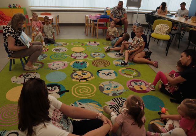 dzieci i rodzice siedzą na kolorowym dywanie, pomiędzy nimi siedzi bibliotekarka z książką. W tle regały z książkami
