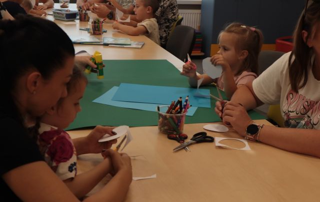 przy połączonych w rząd stołach siedzą dzieci z rodzicami i wycinają i kleją
