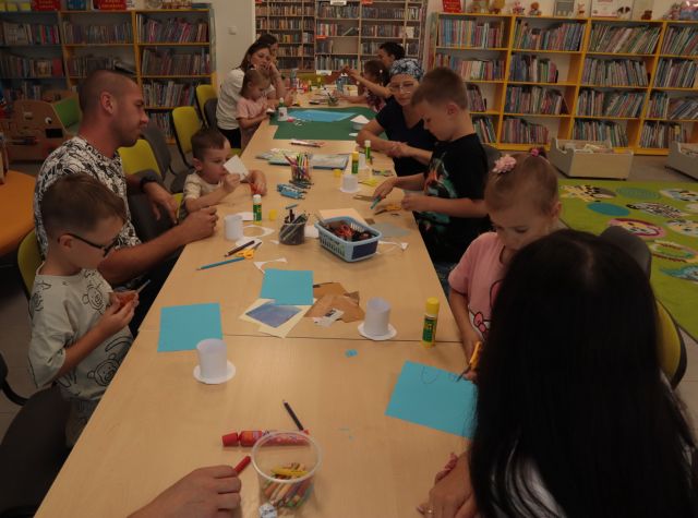 przy połączonych w rząd stołach siedzą dzieci z rodzicami i wycinają i kleją