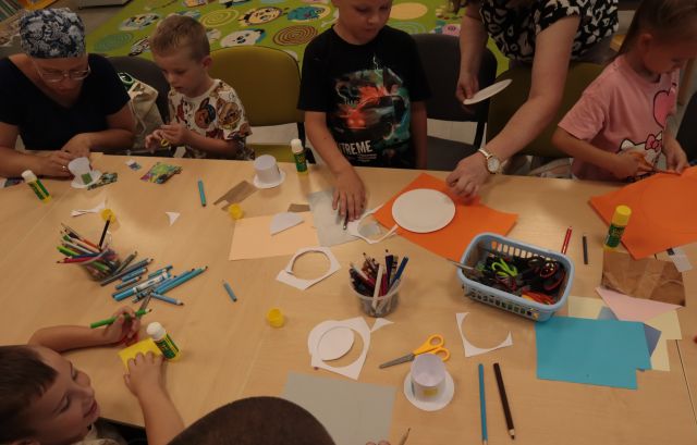 przy połączonych w rząd stołach siedzą dzieci z rodzicami i wycinają i kleją