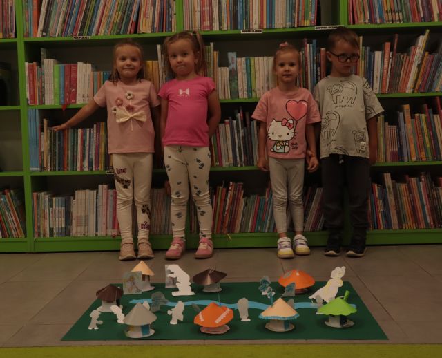 na podłodze leży makieta z wioską smerfów, za nią stoją dzieci, w tle regały z książkami