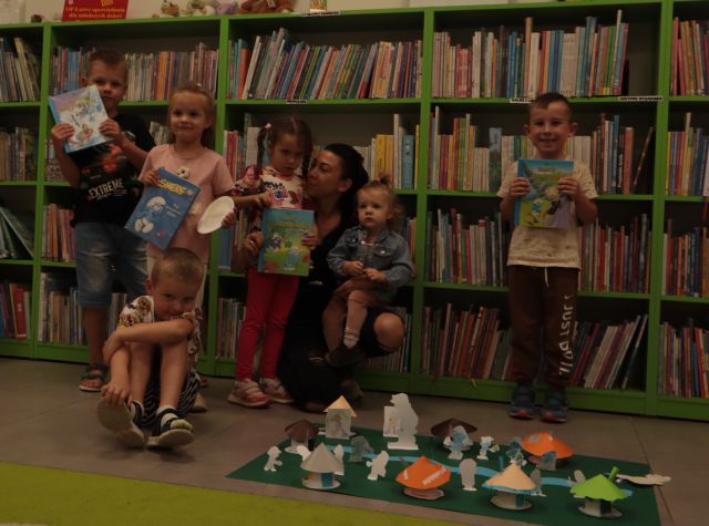 na podłodze leży makieta z wioską smerfów, za nią stoją dzieci, w tle regały z książkami