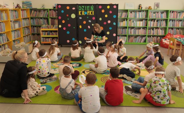 przed bibliotekarką z książką na kolorowym dywanie siedzą dzieci, w tle czarna tablica z naklejonymi kropkami i regały z książkami