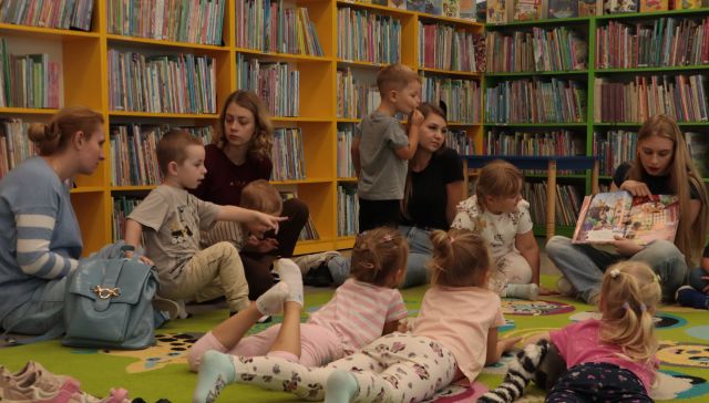 dzieci siedzą i leżą na dywanie, przed nimi siedzi bibliotekarka z książką, w tle regały z książkami