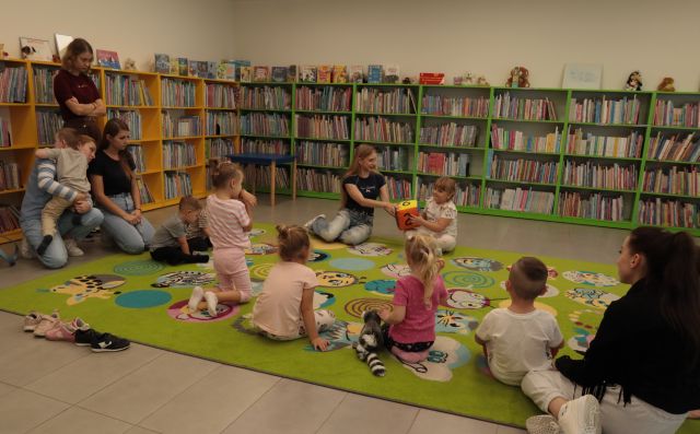 dzieci i bibliotekarką z dużą kolorową kostką siedzą na kolorowym dywanie, w tle regały z książkami