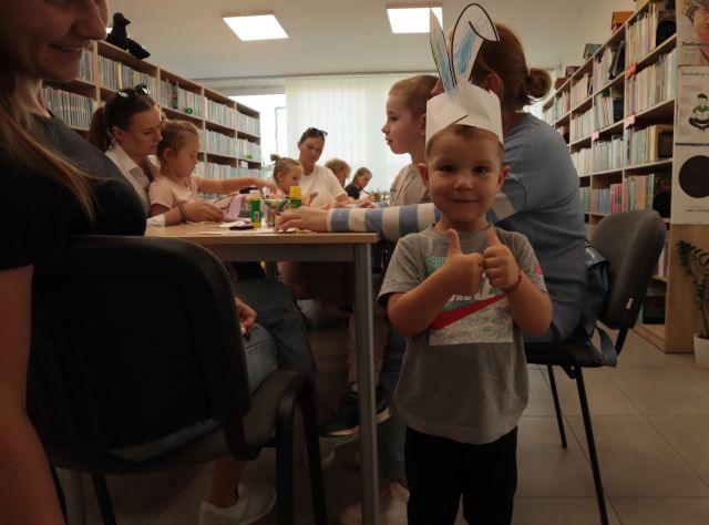 dziecko pozuje do zdjęcia w króliczych uszach wyciętych z papieru, w tle dzieci z rodzicami przy stołach kolorują i wycinają