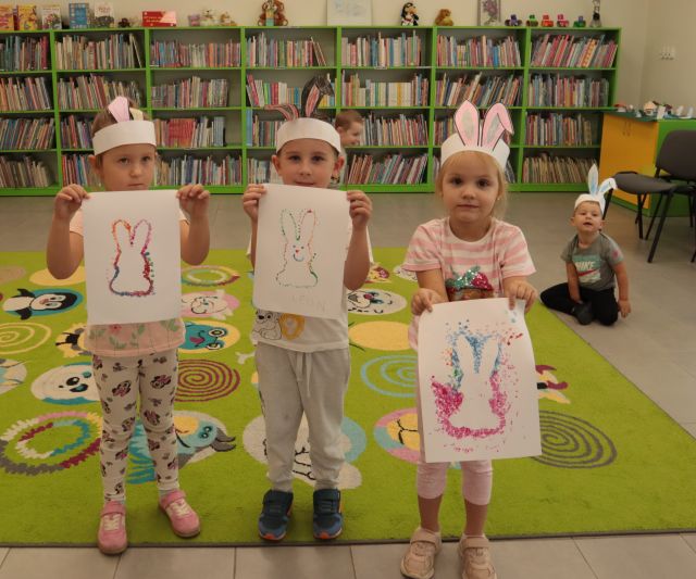 3 dzieci pozuje do zdjęcia z kartkami z namalowanymi królikami i opaskach z króliczymi uszami z papieru na głowach