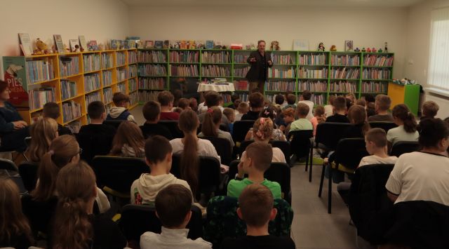 uczniowie siedzą na krzesłach i dywanie, przed nimi stoi autor, w tle regały z książkami