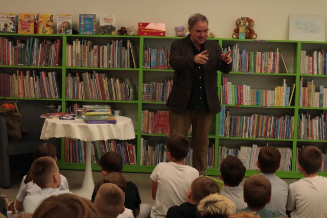 uczniowie siedzą na krzesłach i dywanie, przed nimi stoi autor, w tle regały z książkam