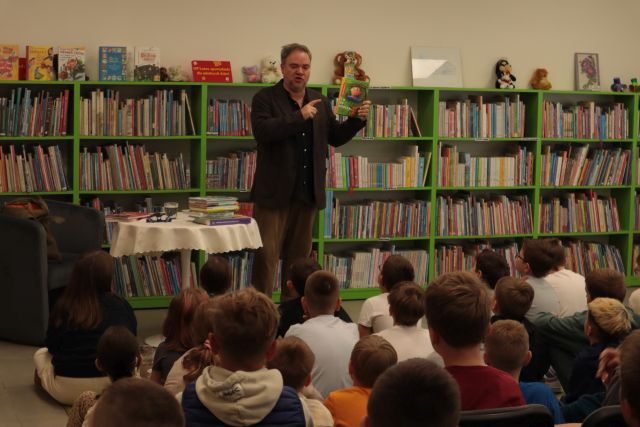 autor z książką stoi przed dziećmi siedzącymi na krzesłach i dywanie