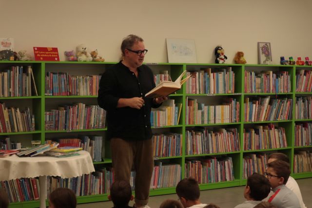 autor z książką stoi przed dziećmi siedzącymi na krzesłach i dywanie