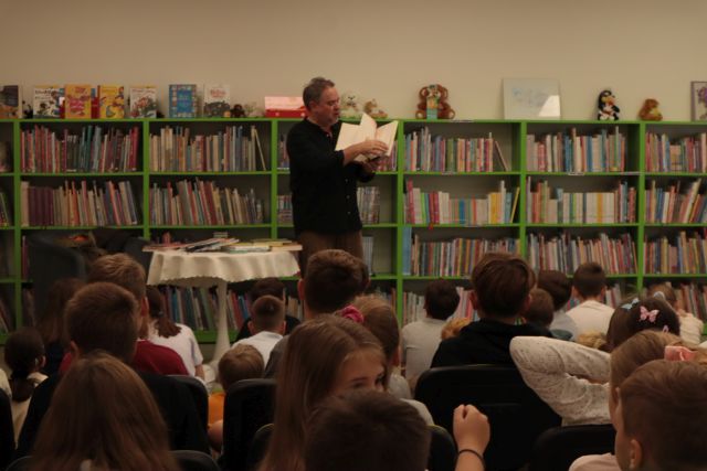 autor z książką stoi przed dziećmi siedzącymi na krzesłach i dywanie