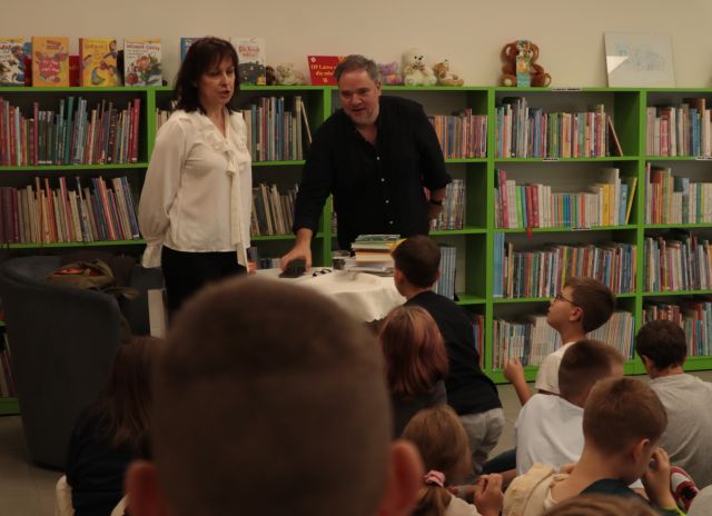 przed dziećmi stoi dyrektorka biblioteki i autor, w tle ragały z książkami