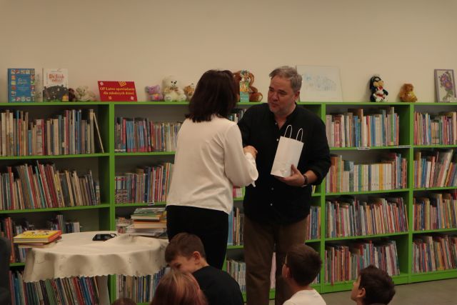 autor z książką stoi przed dziećmi siedzącymi na krzesłach i dywanie